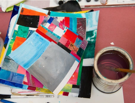 A pile of painted collages next to a bucket of purple-tinged water with a brush in it